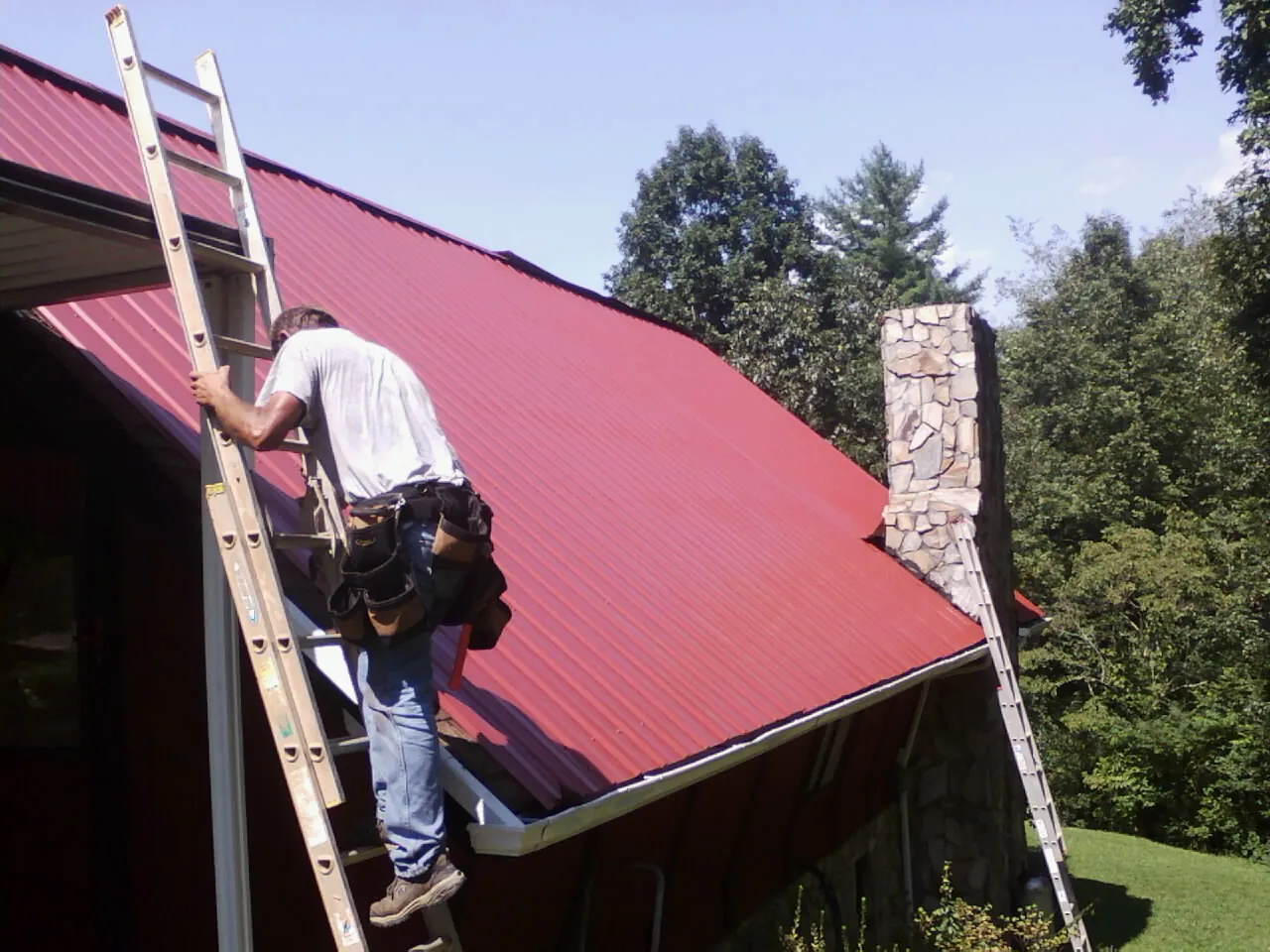 Roofer on a ladder beside a red metal roof and stone chimney — Soffit & Fascia Repair by Roofing Brothers in Shippensburg